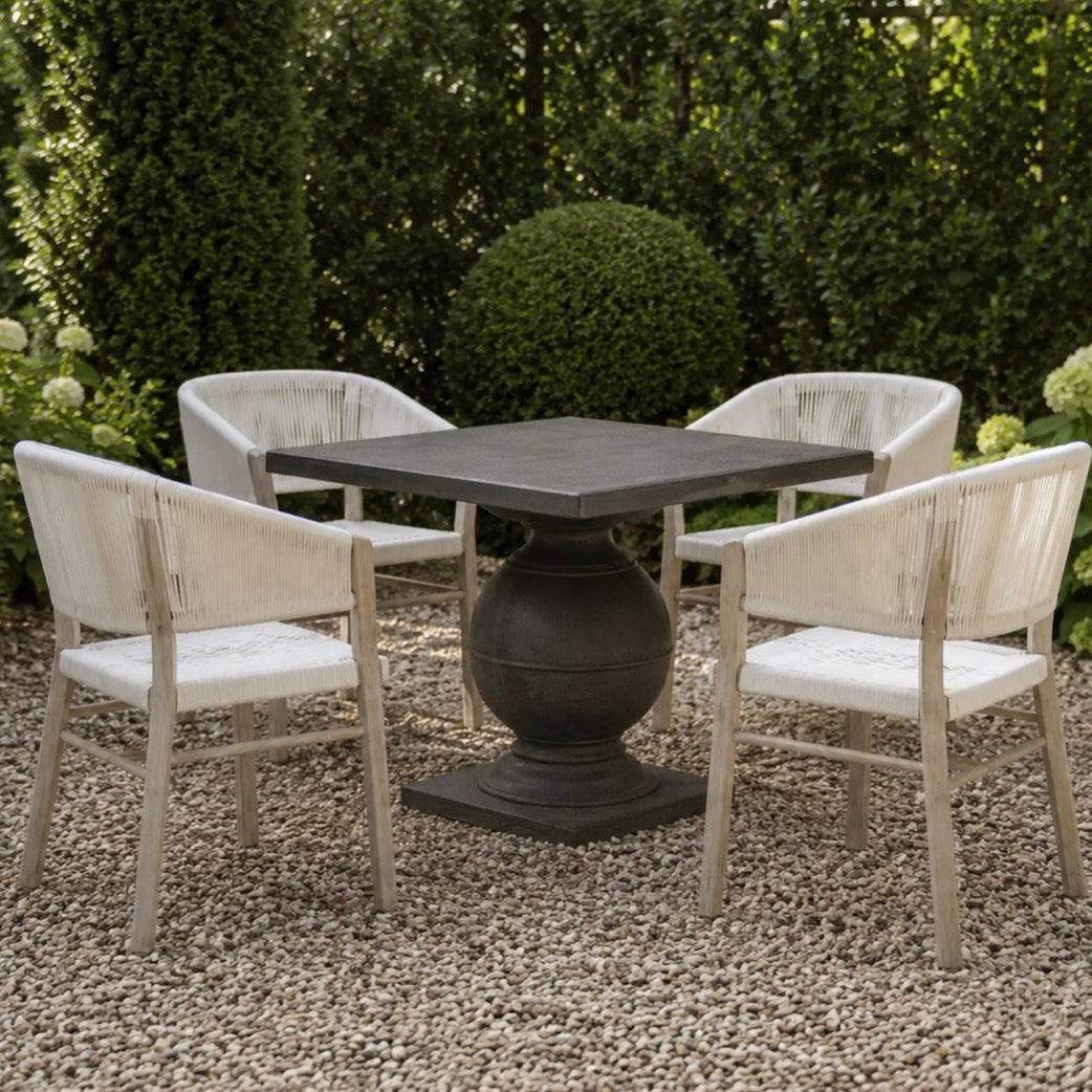 A 4-piece outdoor dining set featuring a square pedestal table and four matching armchairs with distressed off-white frames and white seats, set on a gravel patio in a garden.