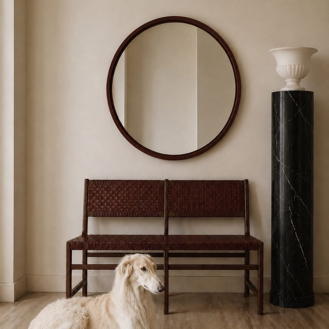 A long-haired cream-colored dog lies on a light wood floor in front of a dark brown woven bench and a round dark wood mirror, with a black marble column and white urn against a beige wall.