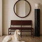 A long-haired cream-colored dog lies on a light wood floor in front of a dark brown woven bench and a round dark wood mirror, with a black marble column and white urn against a beige wall.