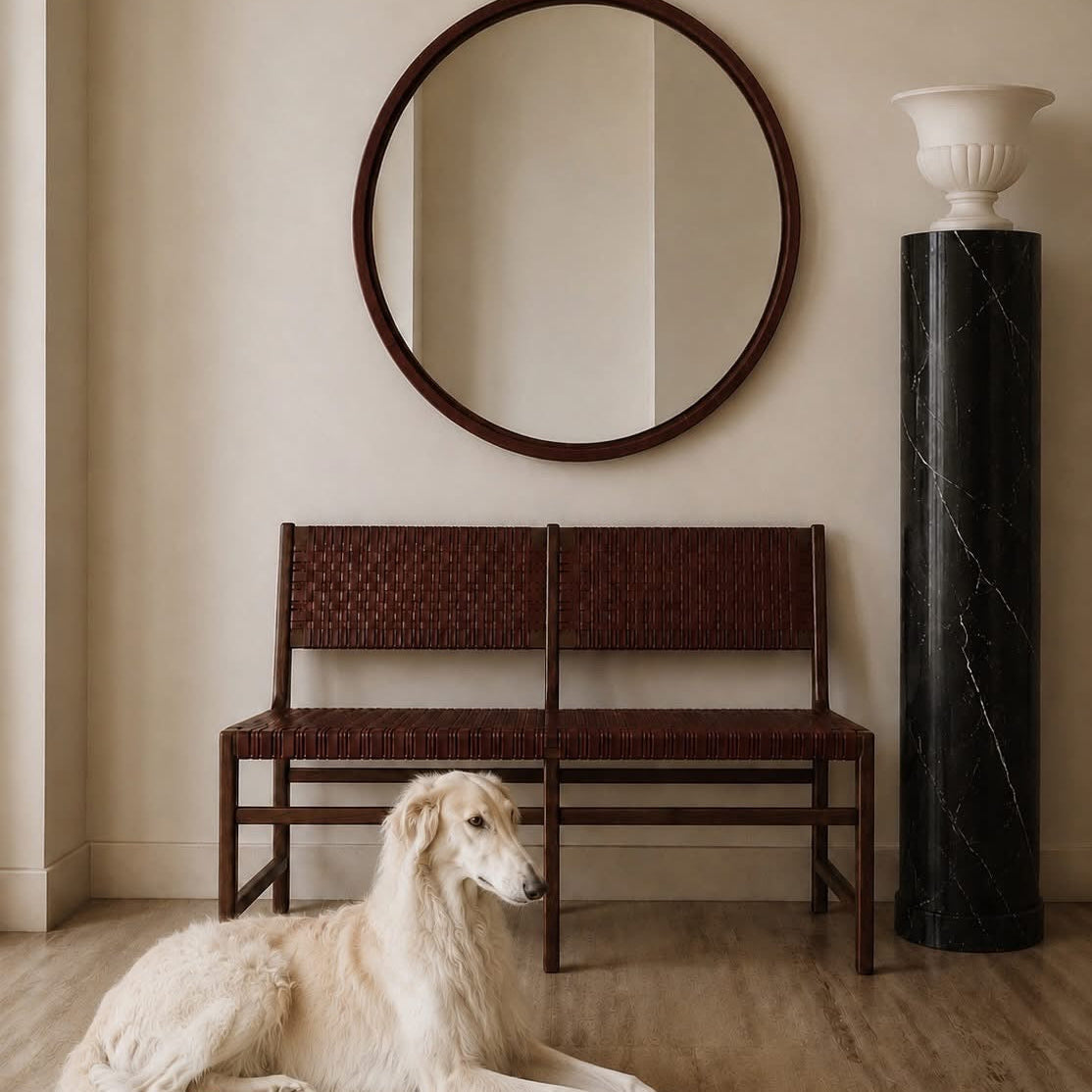 A long-haired cream-colored dog lies on a light wood floor in front of a dark brown woven bench and a round dark wood mirror, with a black marble column and white urn against a beige wall.