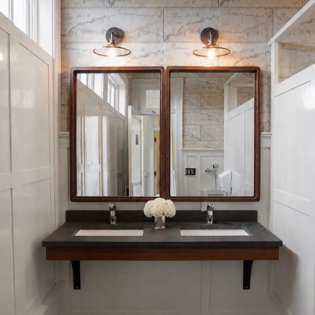 A double-sink bathroom vanity with a dark wood floating countertop, two rectangular mirrors with dark wood frames, and two wall-mounted sconces above, set against a light stone tile wall and white paneled walls.