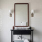 A modern bathroom vanity setup featuring a black console table with a glass shelf, an undermount sink, a large rectangular mirror with a dark wood frame, and two wall-mounted sconces against a white subway tile wall.