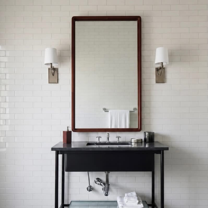 A modern bathroom vanity setup featuring a black console table with a glass shelf, an undermount sink, a large rectangular mirror with a dark wood frame, and two wall-mounted sconces against a white subway tile wall.
