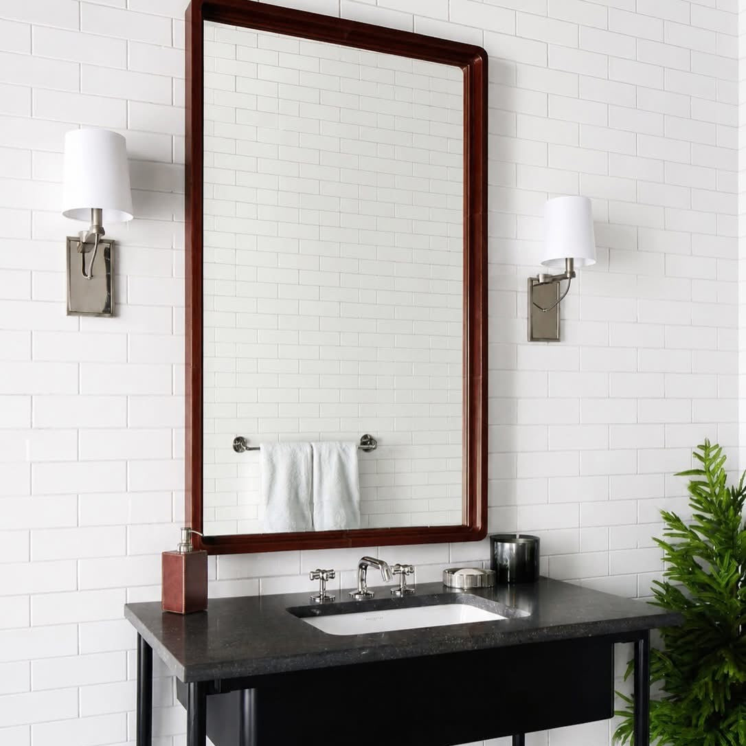 A modern bathroom vanity setup featuring a large rectangular mirror with a dark wood frame, mounted on a white subway tile wall. Below the mirror is a black floating vanity cabinet with a dark stone countertop, integrated white sink, chrome faucet, and exposed silver plumbing.