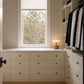 A walk-in closet with cream-colored built-in drawers and shelving, a window with a beige roller shade, a small table lamp on the counter, and hanging black-and-white striped garments.