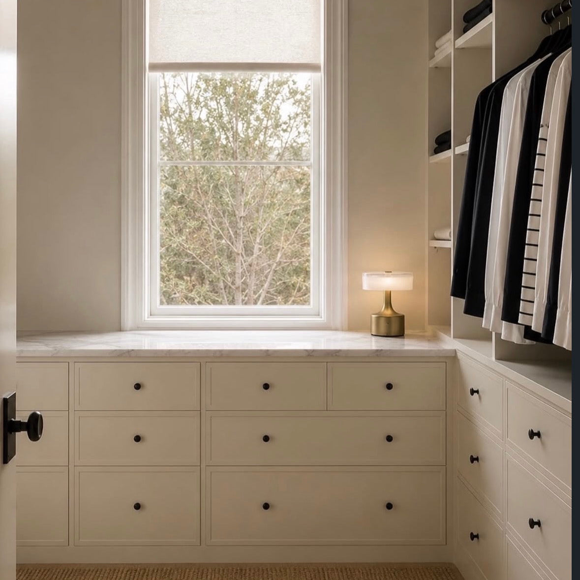A walk-in closet with cream-colored built-in drawers and shelving, a window with a beige roller shade, a small table lamp on the counter, and hanging black-and-white striped garments.