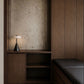 A modern interior corner featuring a dark wood built-in cabinet and bench with a textured beige back panel and a lit table lamp on the shelf.