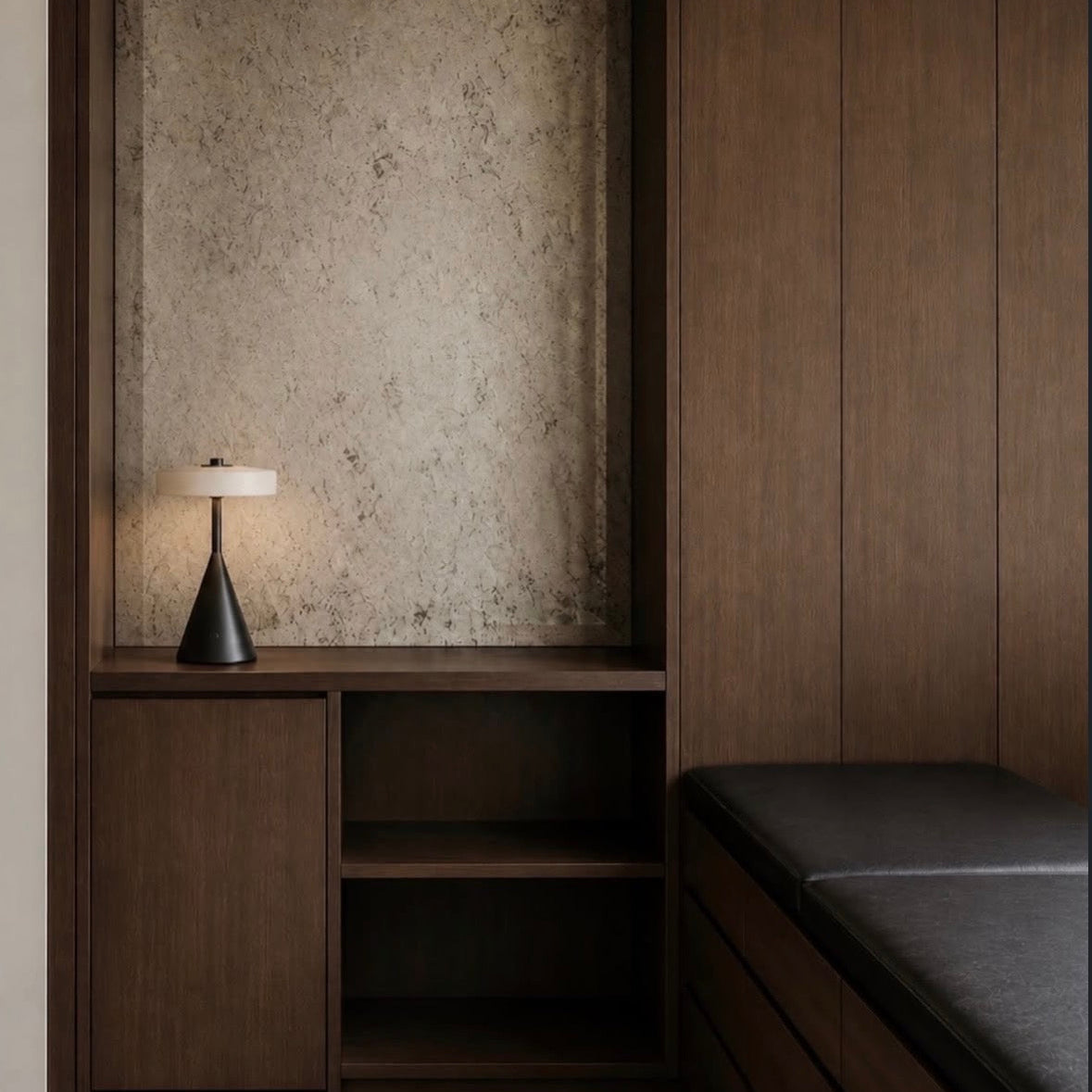 A modern interior corner featuring a dark wood built-in cabinet and bench with a textured beige back panel and a lit table lamp on the shelf.
