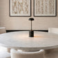 A modern dining room scene featuring a round white marble table with a black marble base and a black dome lamp, surrounded by cream upholstered chairs. Two framed abstract chain prints hang on a textured beige wall above the table.