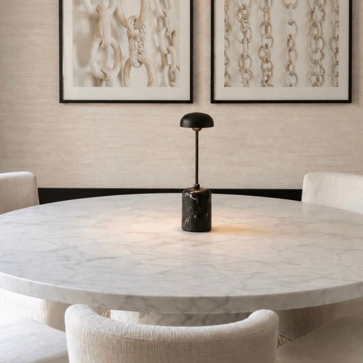 A modern dining room scene featuring a round white marble table with a black marble base and a black dome lamp, surrounded by cream upholstered chairs. Two framed abstract chain prints hang on a textured beige wall above the table.