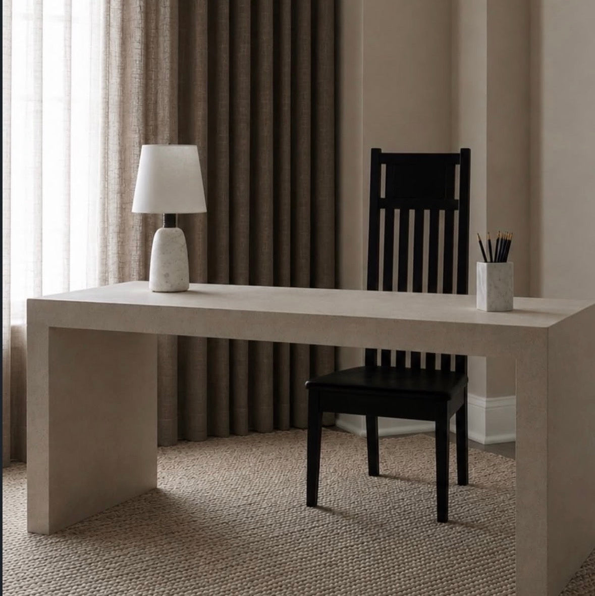 A minimalist home office scene featuring a long, low concrete desk, a black wooden slat-back chair, a white table lamp, and a small white cup with pens, set against a background of beige walls and dark brown curtains.