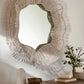 An organic-shaped wall mirror with a wide, sculptural frame made of many thin, light-colored sticks radiating outward, mounted on a beige wall above a styled tabletop with a vase, bowl, and small plant.