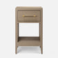 A modern bedside table with a single drawer and open lower shelf in a warm taupe-gray finish, featuring gold-finished hardware. It is styled in a bedroom next to a bed with white linens, a beige upholstered headboard, a wall-mounted brass sconce, and a patterned rug.