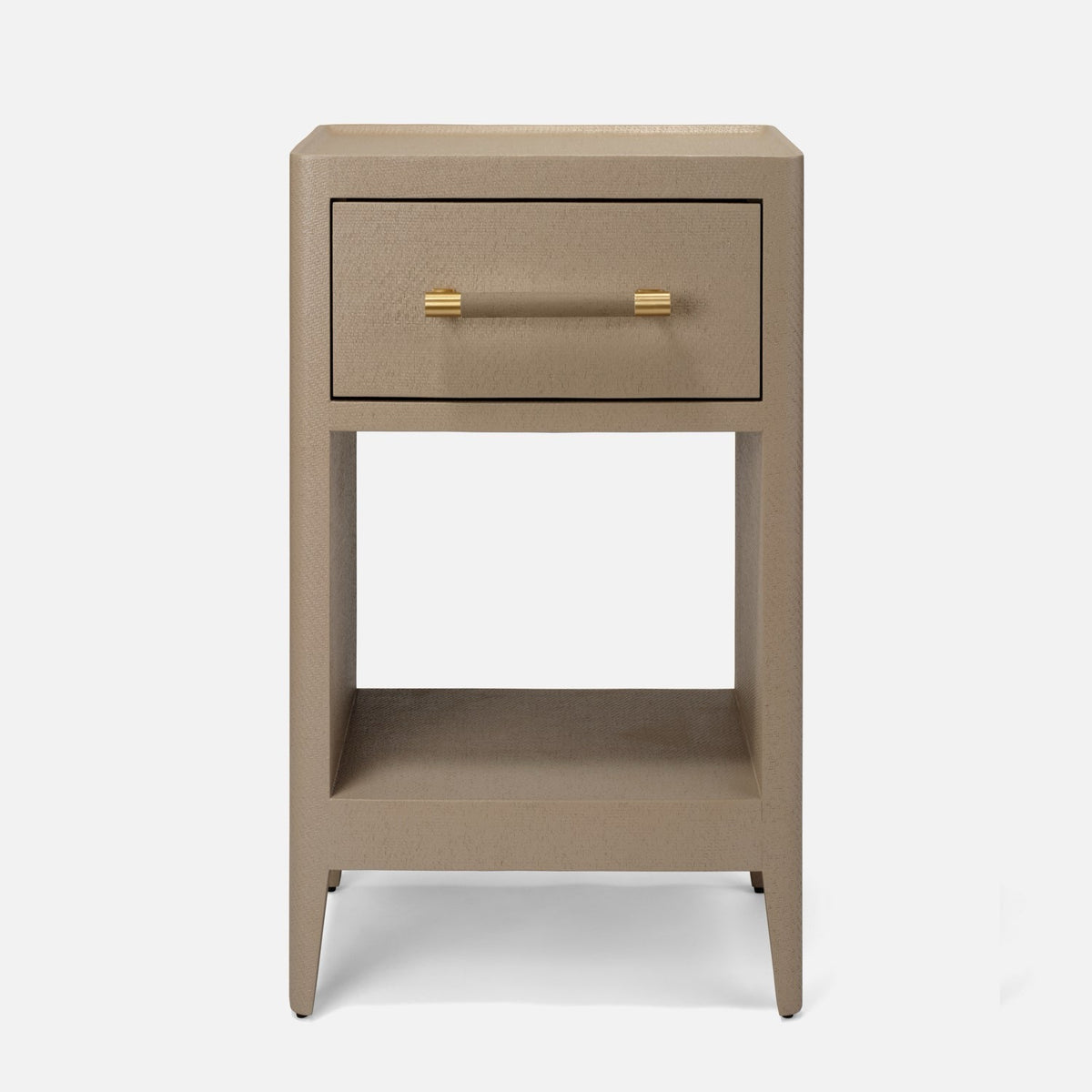 A modern bedside table with a single drawer and open lower shelf in a warm taupe-gray finish, featuring gold-finished hardware. It is styled in a bedroom next to a bed with white linens, a beige upholstered headboard, a wall-mounted brass sconce, and a patterned rug.
