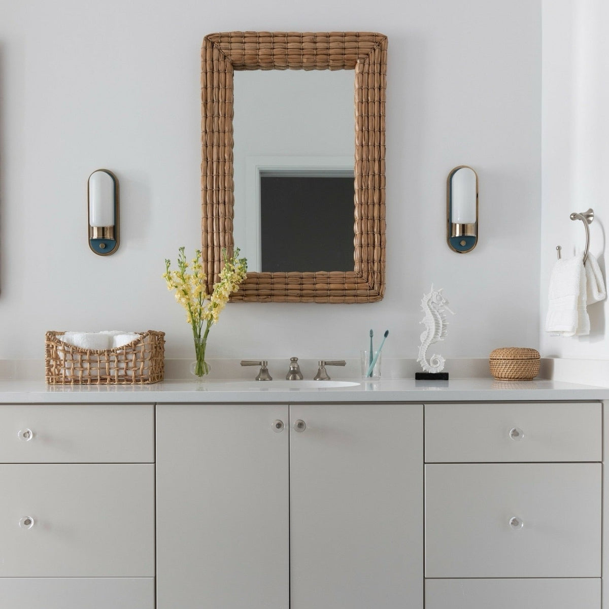 A modern bathroom vanity setup featuring a light gray double-sink cabinet with a white countertop, a large rectangular wicker mirror, blue and white patterned floor tiles, and matching wicker decorative baskets.