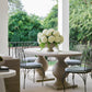 An outdoor dining scene on a covered patio with a round stone pedestal table, black metal chairs with light blue cushions, and pink hydrangea bouquets in the center, with lush green trees in the background.