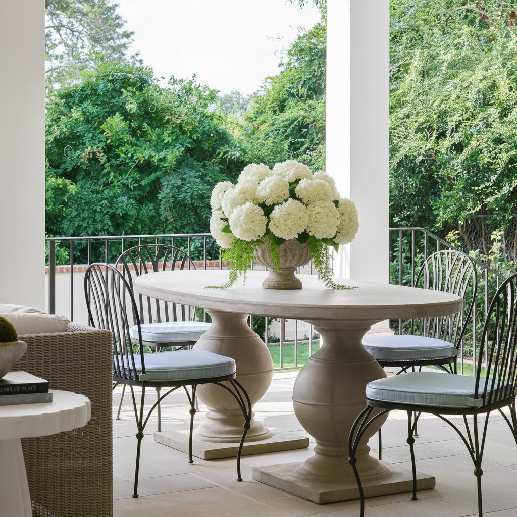 An outdoor dining scene on a covered patio with a round stone pedestal table, black metal chairs with light blue cushions, and pink hydrangea bouquets in the center, with lush green trees in the background.