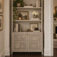 A tall built-in shelving unit with a textured light gray wood frame and two cabinet doors, styled with books, bowls, a potted plant, and framed art in a home entryway.
