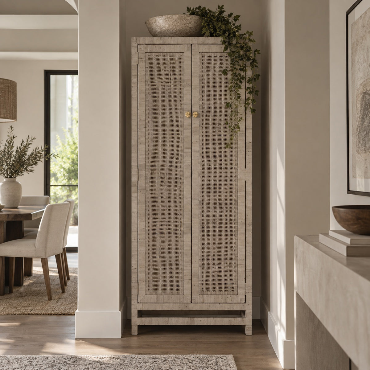 A tall two-door cabinet with woven cane or rattan panels and small gold knobs, shown in a real interior hallway with a dining area on the left and a console table on the right.