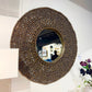 A large round decorative wall mirror with a wide, textured frame made of many small brown beads, mounted on a white wall. The mirror reflects an indoor room, and a white lamp and flowers are partially visible in the foreground.