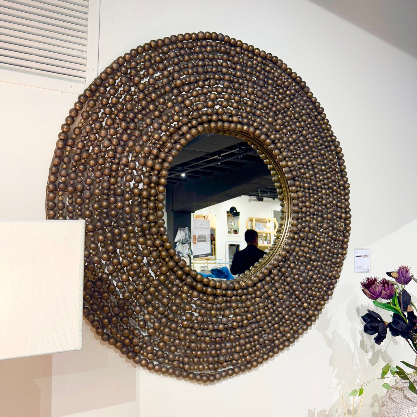 A large round decorative wall mirror with a wide, textured frame made of many small brown beads, mounted on a white wall. The mirror reflects an indoor room, and a white lamp and flowers are partially visible in the foreground.