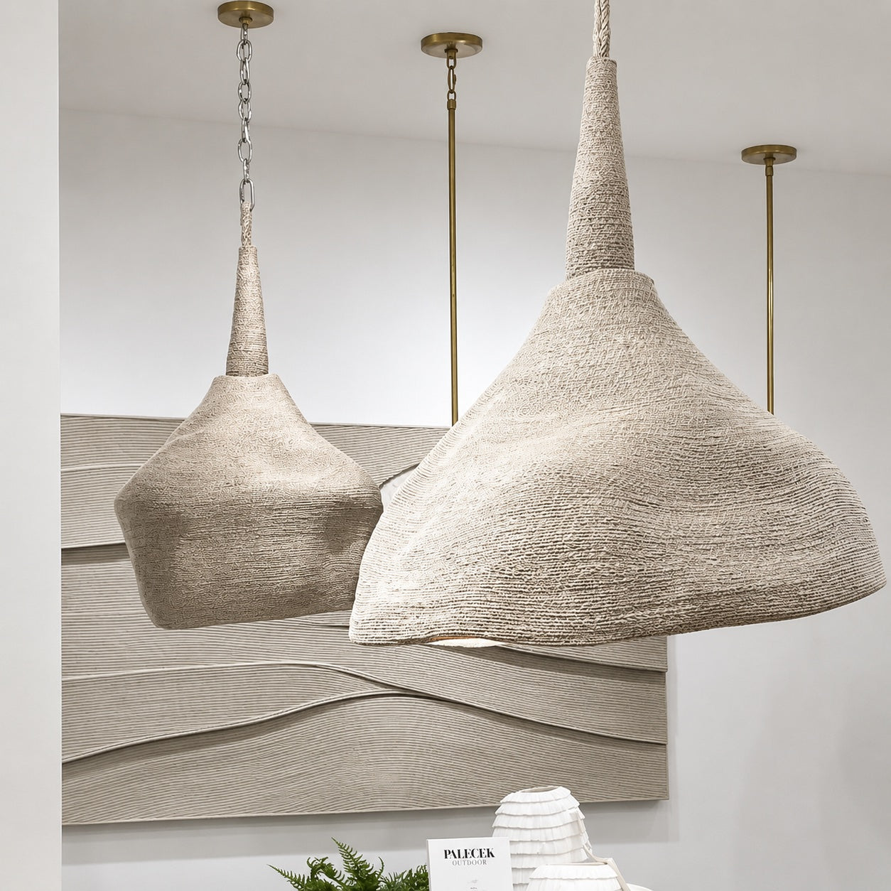 Two woven pendant light shades in a textured beige material hang from brass rods against a white wall, with a decorative abstract panel behind them.