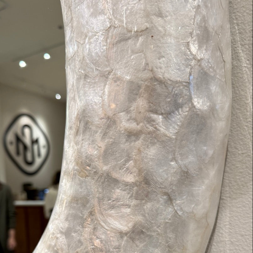 A close-up of a large, organically shaped mirror with a textured, pebble-like surface in off-white and light gray tones, mounted on a textured wall. The background is a blurred interior shop setting with a logo on the wall.