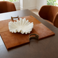 A white sculptural leaf-shaped bowl sits on a brown leather placemat on a dark wood dining table, with two metallic gold curved coasters and brown upholstered chairs in a bright room.