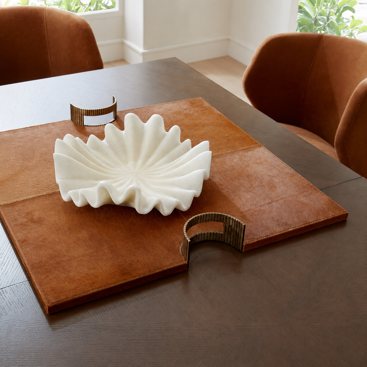 A white sculptural leaf-shaped bowl sits on a brown leather placemat on a dark wood dining table, with two metallic gold curved coasters and brown upholstered chairs in a bright room.