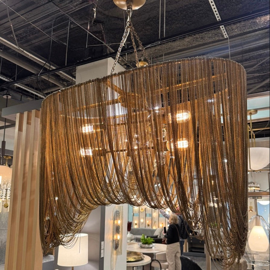 Large gold chandelier hanging in a showroom with people and other lighting fixtures in the background.