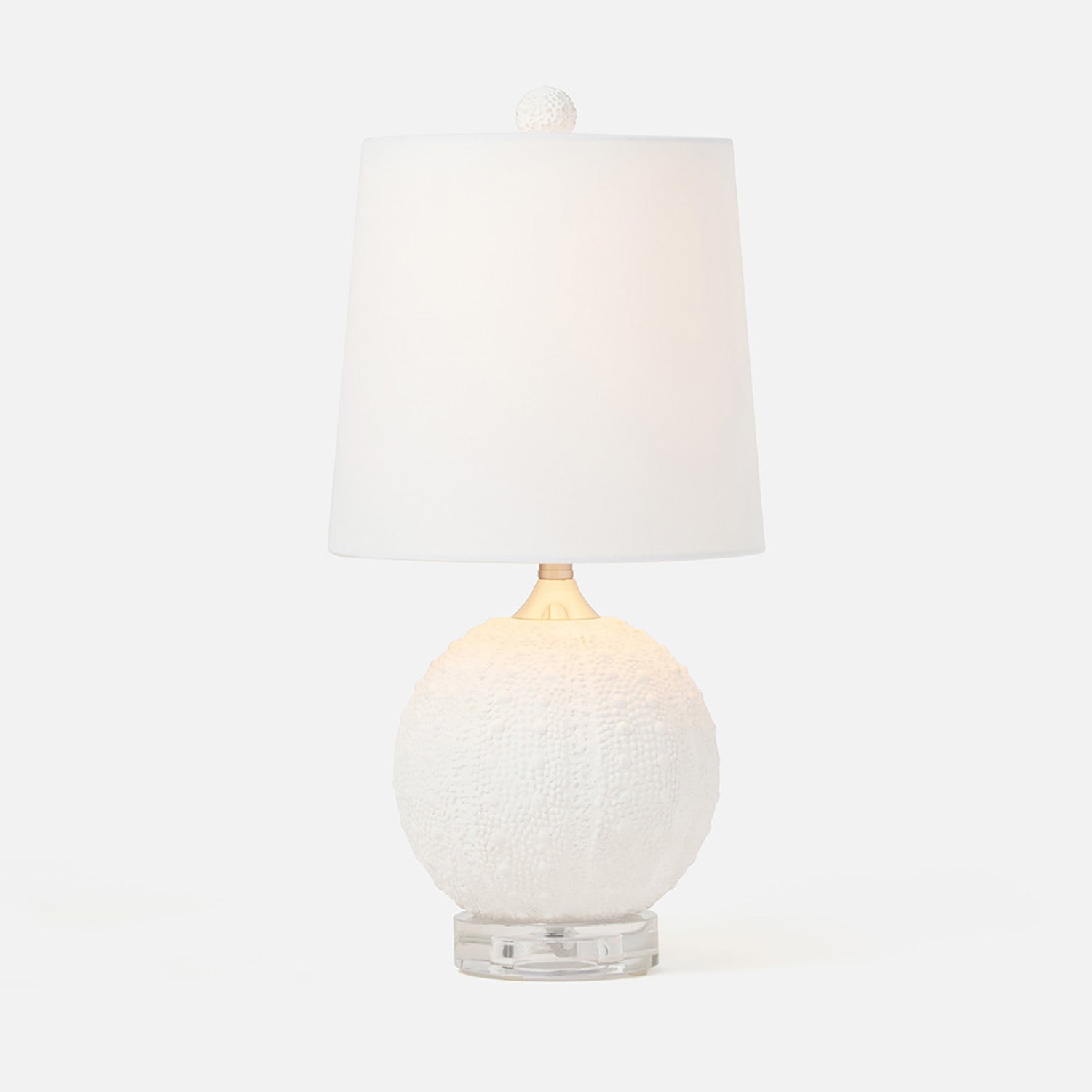 A table lamp with a textured white spherical base, a clear acrylic pedestal foot, and a white fabric drum shade with a small white finial on top, photographed against a clean white background.