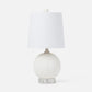 A table lamp with a textured white spherical base, a clear acrylic pedestal foot, and a white fabric drum shade topped with a small white pom-pom.