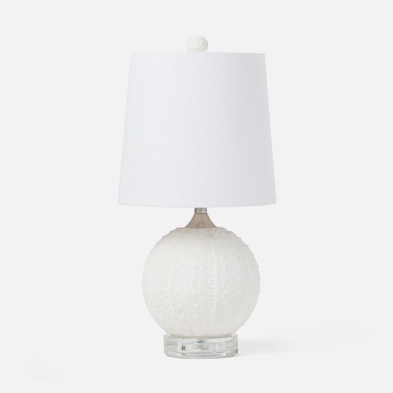 A table lamp with a textured white spherical base, a clear acrylic pedestal foot, and a white fabric drum shade topped with a small white pom-pom.