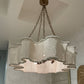 A large multi-light chandelier with a scalloped, wavy white woven shade and bronze-toned hanging hardware, shown hanging in a decorated showroom with wallpaper, a mirror, and a white sculptural floor lamp.