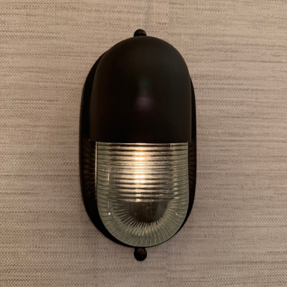 A black oval wall sconce with a ribbed clear glass shade, mounted on a textured beige wall.