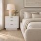 A modern white nightstand with horizontal grooved drawers and gold-tone handles, styled in a bedroom next to a bed with neutral linens and a lamp.