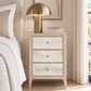 A three-drawer nightstand in a light natural wood finish with distressed white drawer fronts and small brass knobs, styled in a bedroom next to a bed with a gold dome lamp on top.