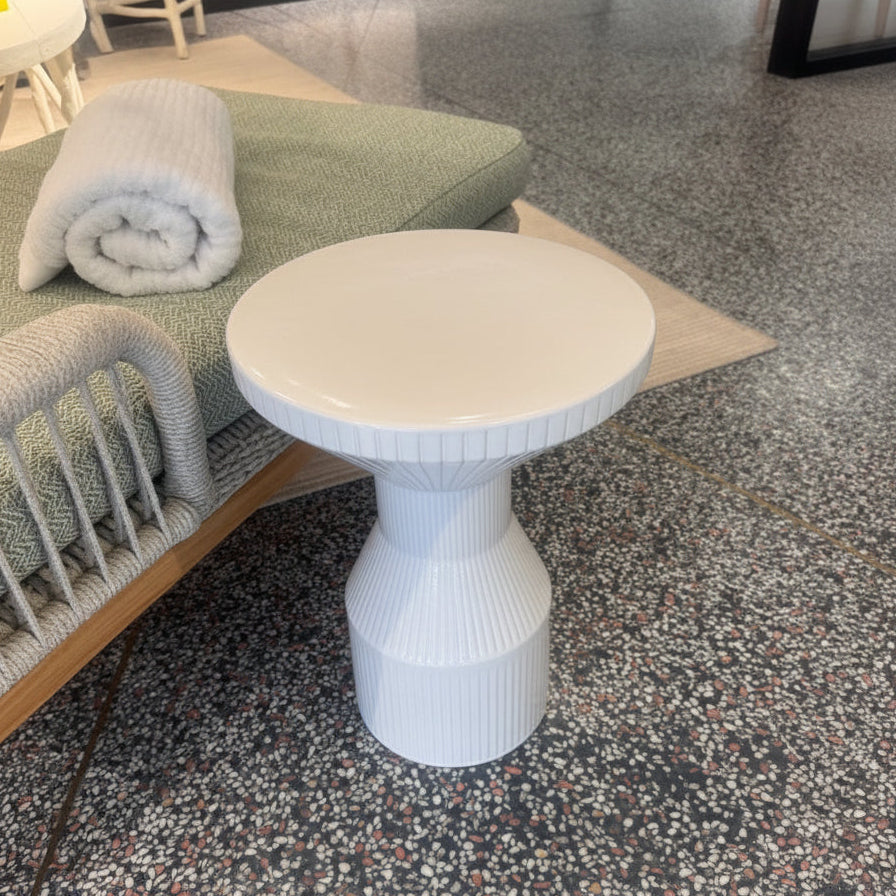 A round white side table with a ribbed pedestal base, shown in a furniture showroom next to a green cushioned bench on a speckled terrazzo floor.