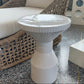 A white ribbed pedestal side table with a round top, positioned in front of a woven rattan armchair with blue and white striped cushions on a speckled terrazzo floor.