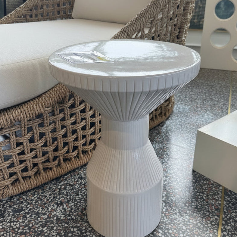 A white ribbed pedestal side table with a round top, positioned in front of a woven rattan armchair with blue and white striped cushions on a speckled terrazzo floor.