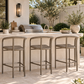 Three outdoor bar stools with metal frames and woven seats are positioned at a stone kitchen island in a backyard patio setting with a grill, plants, and a fence in the background.