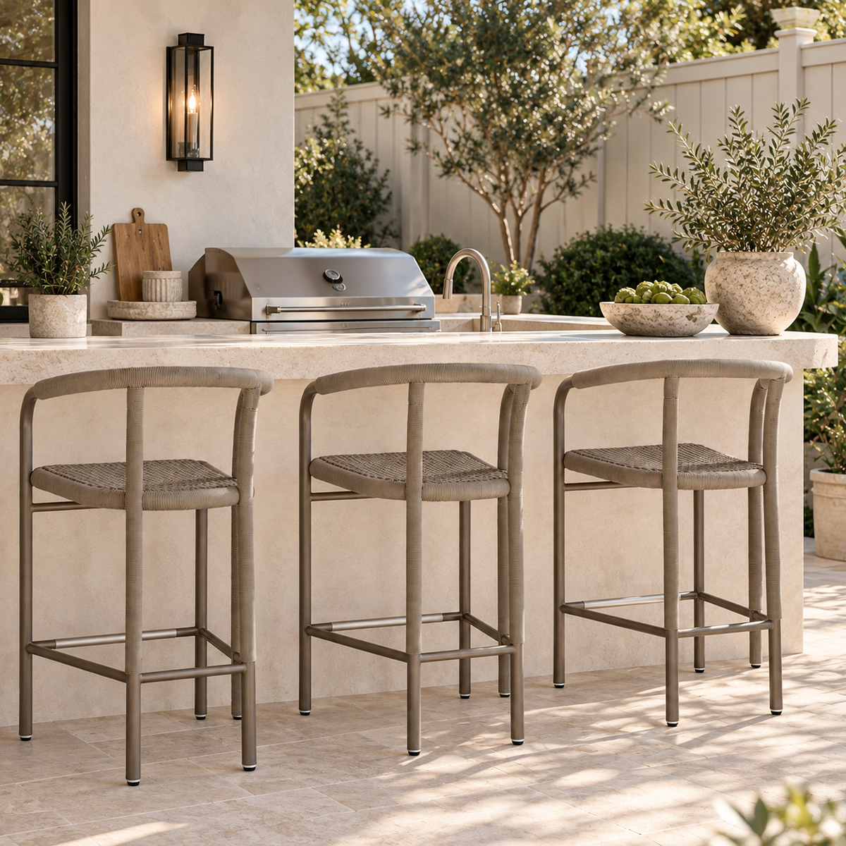 Three outdoor bar stools with metal frames and woven seats are positioned at a stone kitchen island in a backyard patio setting with a grill, plants, and a fence in the background.