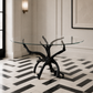 A round glass-top table with a sculptural black branch-like base, placed in a styled interior with cream walls, a black-and-white geometric tile floor, and wall sconces.