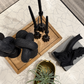 A styled tabletop arrangement featuring black decorative sculptural objects, a wooden tray, a gold planter with a green air plant, and a gray base with a black branch-like decor piece, all on a white cracked-geometric surface.