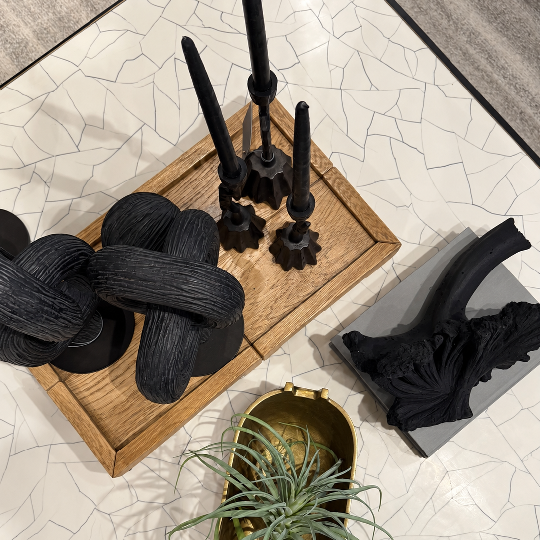 A styled tabletop arrangement featuring black decorative sculptural objects, a wooden tray, a gold planter with a green air plant, and a gray base with a black branch-like decor piece, all on a white cracked-geometric surface.