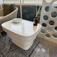 A modern white ceramic side table with a glossy tiled top and a ribbed pedestal base, styled in a cafe or lounge setting next to a seated person.