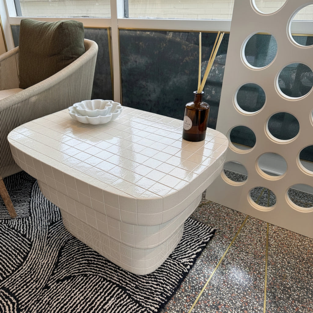 A modern white ceramic side table with a glossy tiled top and a ribbed pedestal base, styled in a cafe or lounge setting next to a seated person.