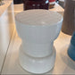 A white ceramic barrel-style stool with a quilted top and ribbed body, displayed on a white shelf in a furniture store alongside other home decor items.