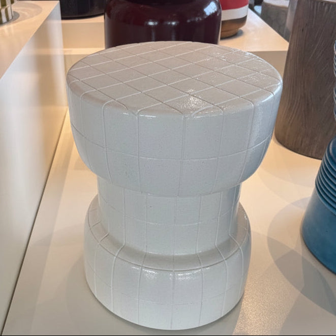 A white ceramic barrel-style stool with a quilted top and ribbed body, displayed on a white shelf in a furniture store alongside other home decor items.