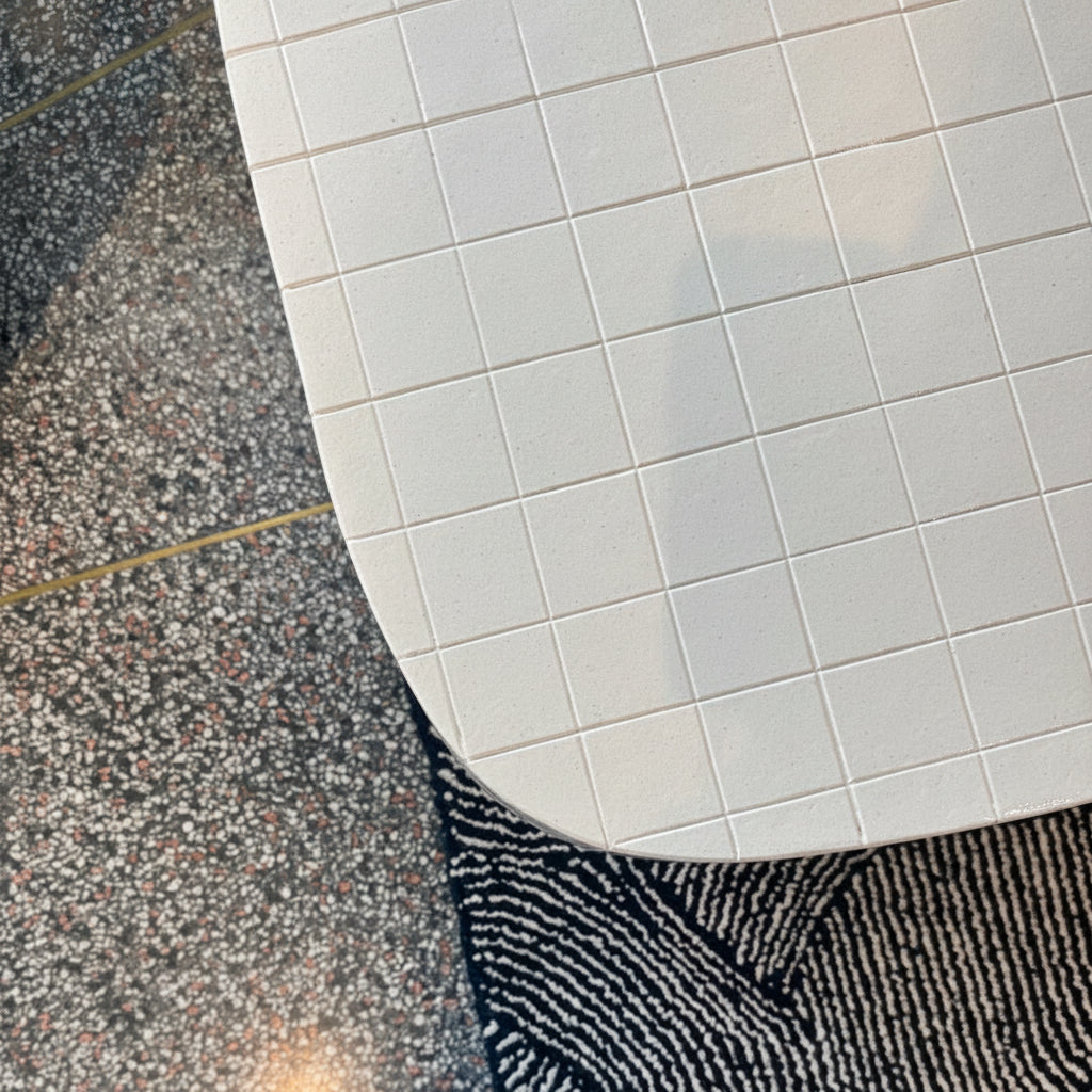 A top-down view of a person's feet wearing brown suede sneakers with white laces, blue jeans, standing on a black and white patterned rug next to a white tiled table on a speckled gray floor.